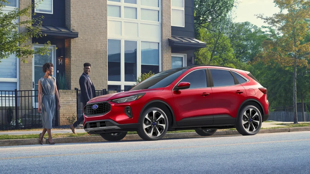 Red 2025 Ford Escape ST-Line Hybrid SUV driving past greenery and buildings in an urban setting.