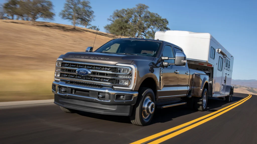 Brown 2025 Ford Super Duty F-350 towing a large trailer on a highway through open countryside.