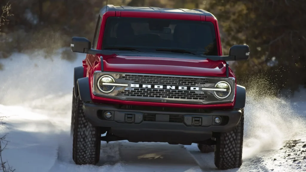 Red 2025 Bronco® SUV driving offroad through snowy terrain, headlights on and snow spraying.