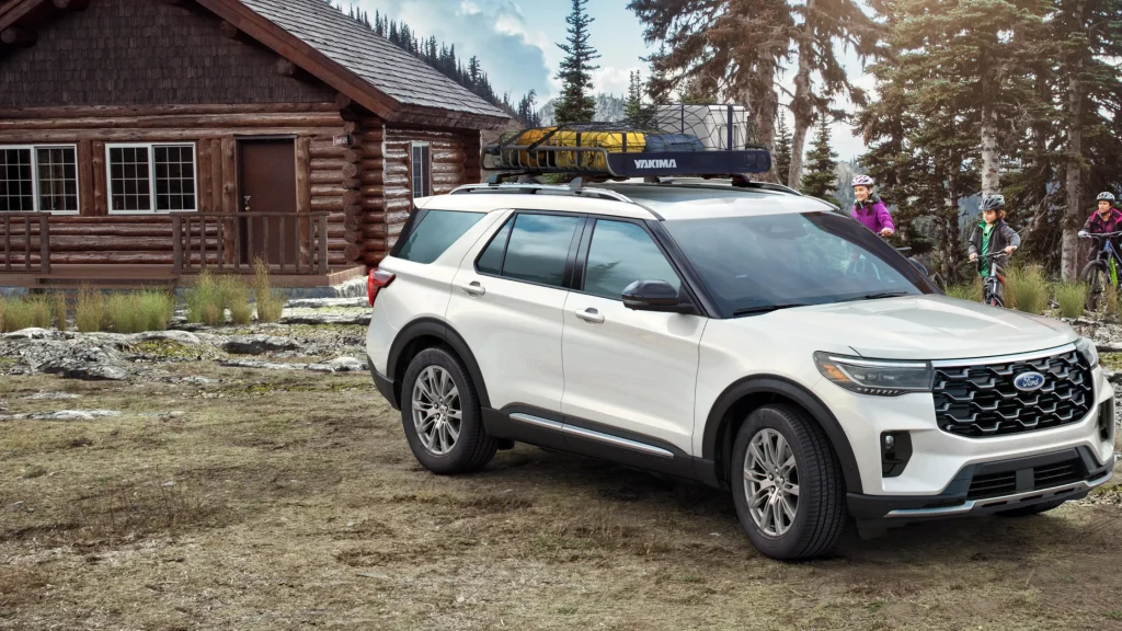 White 2025 Explorer® SUV parked near log cabin with roof rack and family cycling in mountains.