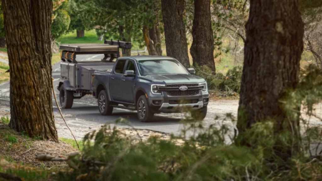 Gray 2025 Ranger® pickup towing adventure trailer through wooded road, framed by tall trees.