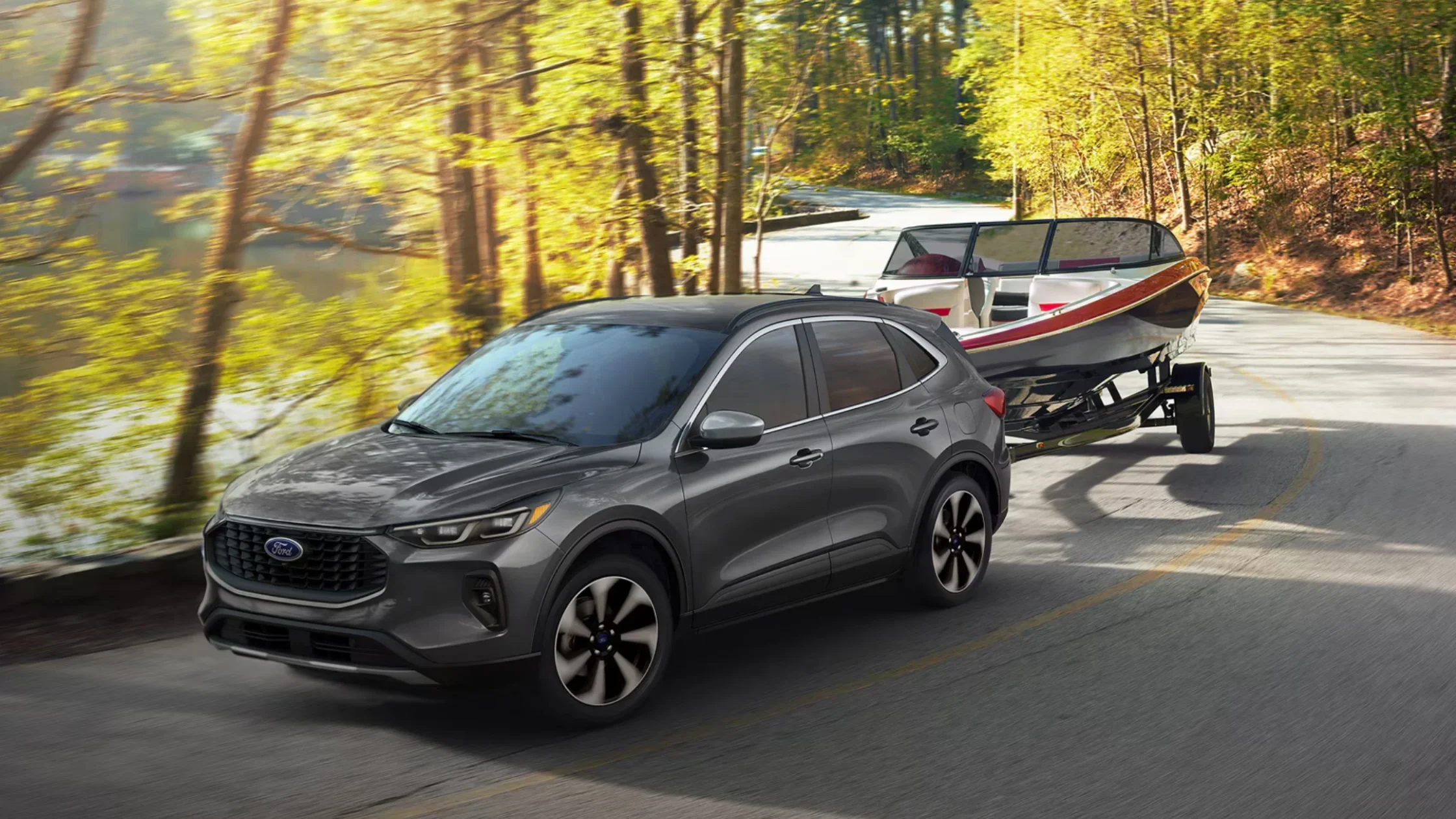 Gray 2025 Escape® SUV towing a boat on a winding forest road near a lake.