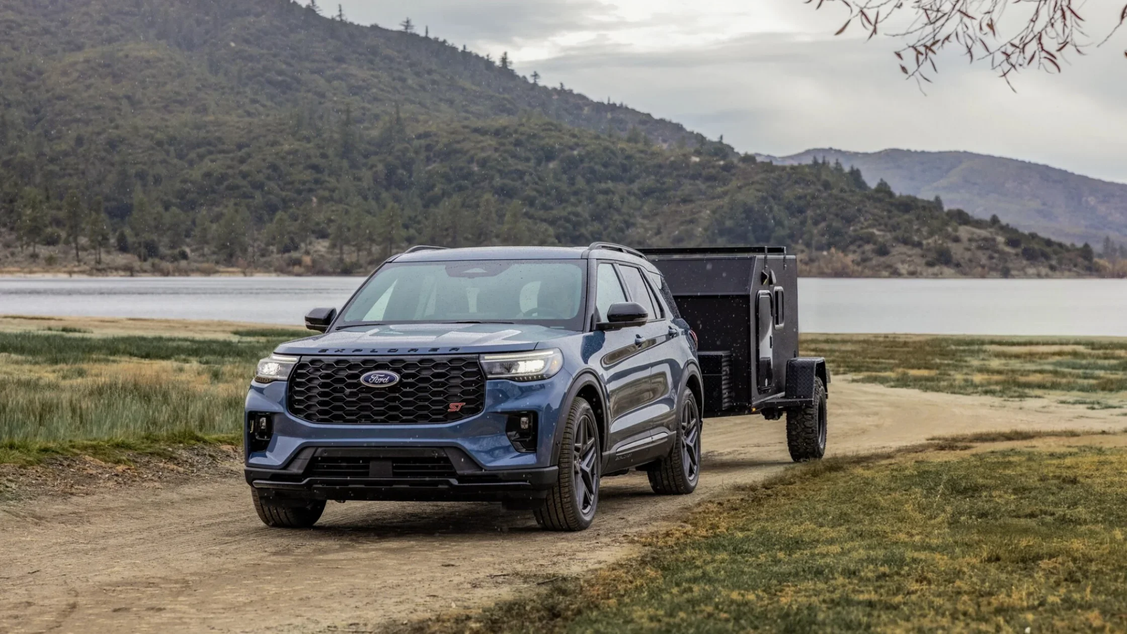 Blue 2025 Explorer® ST towing black trailer on dirt road near mountain lake.