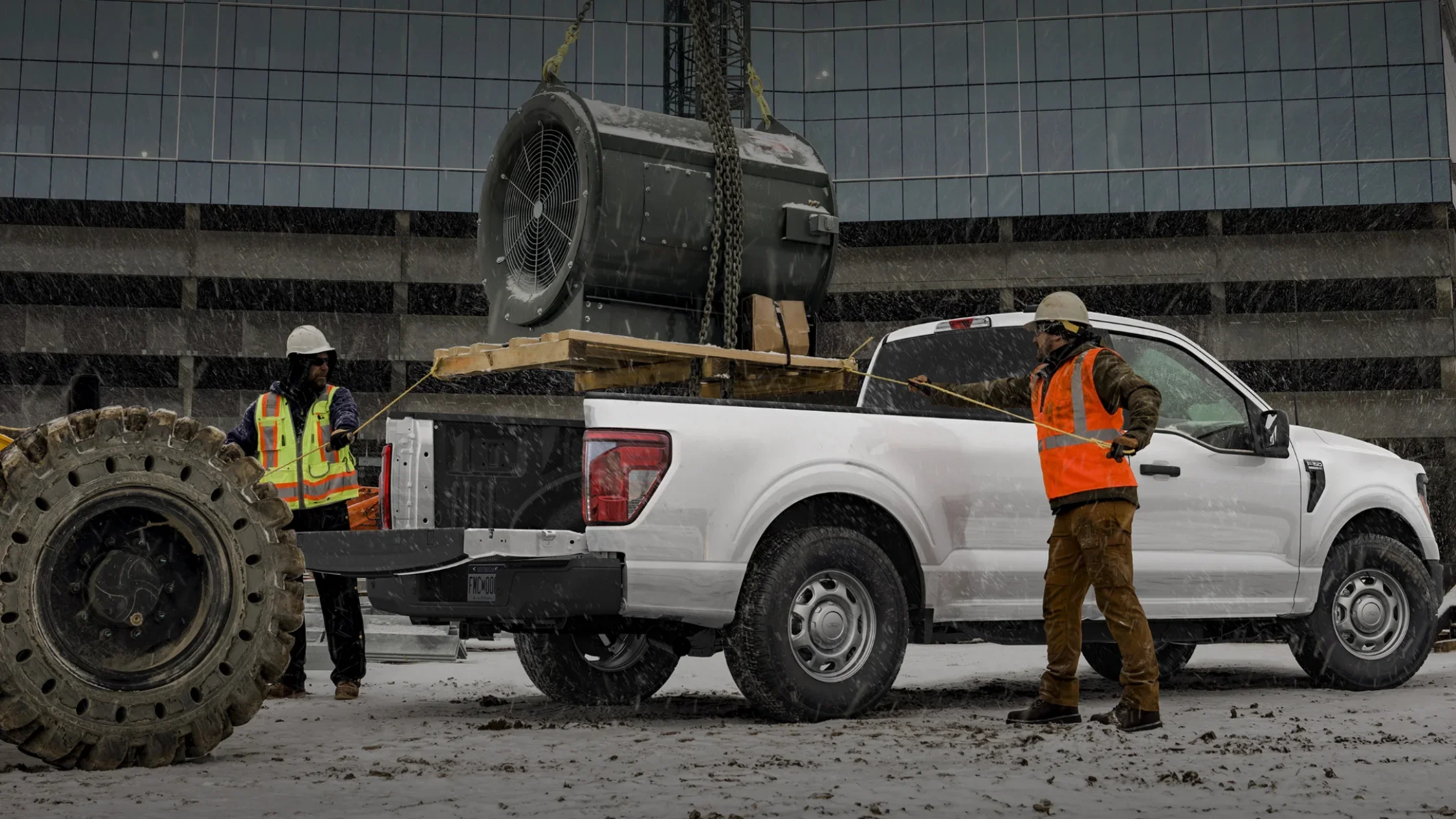 2025 F-150® truck at worksite with crew loading heavy machinery in snowy weather.
