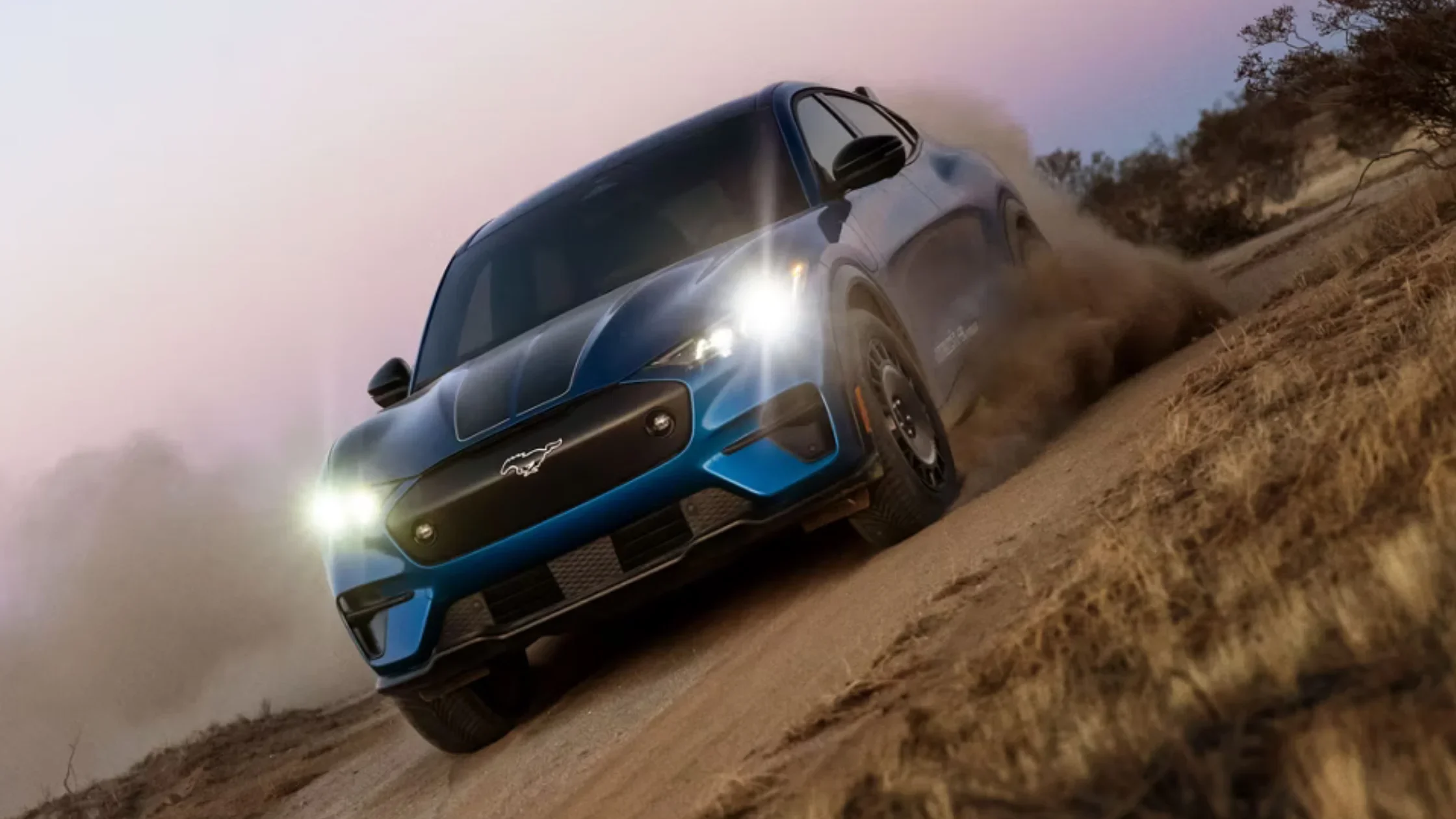 2025 Mustang Mach-E® electric SUV driving on dirt with headlights on at dusk desert road